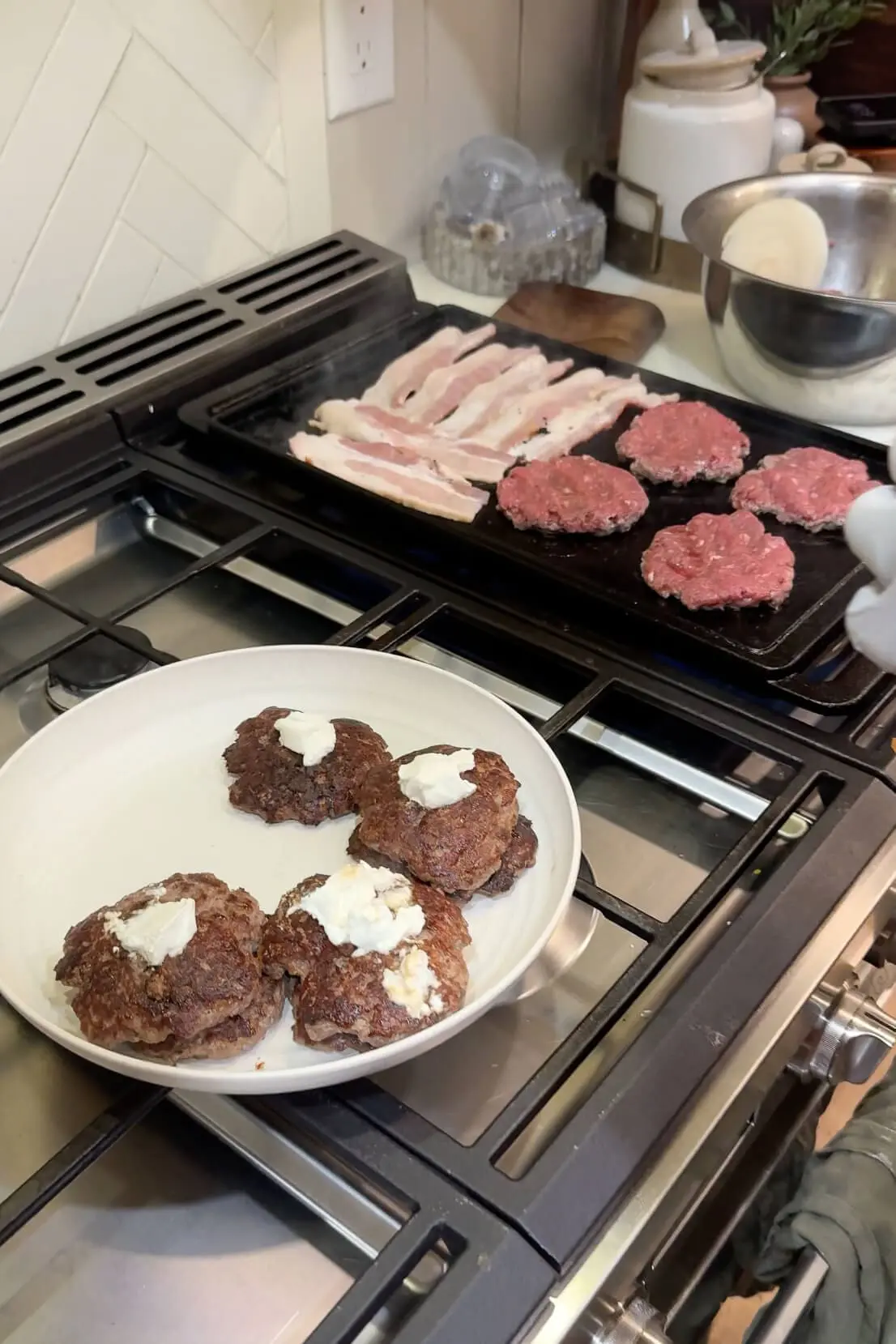 A stovetop griddle loaded with sizzling bacon strips and burger patties cooking to perfection. In the foreground, cooked patties are resting on a plate, each topped with a dollop of creamy goat cheese.